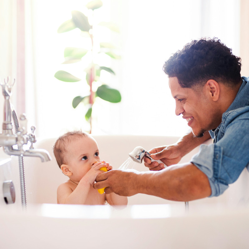 A dad giving his child a bath. Both are smiling. There is a yellow rubber duck too.